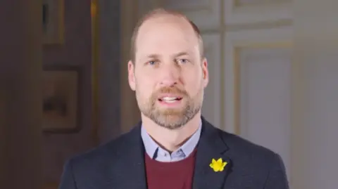 Prince William looking at the camera. He has a daffodil on his left lapel of his blue jacket. 