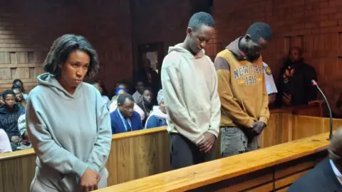 A woman, in a grey hoodie, and two men are seen standing in a courtroom where they face a murder charge