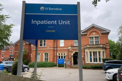A sign reading St Barnabas Inpatient Unit, Lincoln, stands in front of a brick building with cars parked in front of it