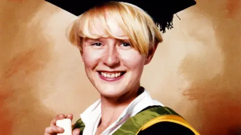 A graduation picture of Melanie Hall, wearing a white collared shirt under green and yellow robes with a mortar board. She has short blonde hair and blue eyes, and is smiling at the camera while holding a scroll. The background is various hues of orange.