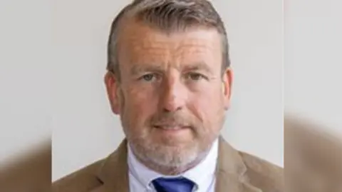 A head and shoulders profile picture of councillor David Maclean, who has short grey hair and beard, is wearing a brown suit, white shirt and blue tie, against a white background