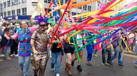 A parade of people who are dressed in bright colours and sequins. They are holding multi-coloured pieces of fabric.