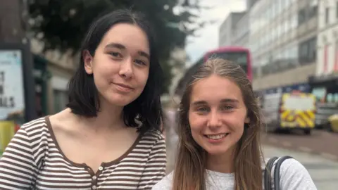 Two teenage girls. the one on the left has a black bob, and a stripey top. The one on the right has long light brown hair, a whit t shirt and a shoulder bag,