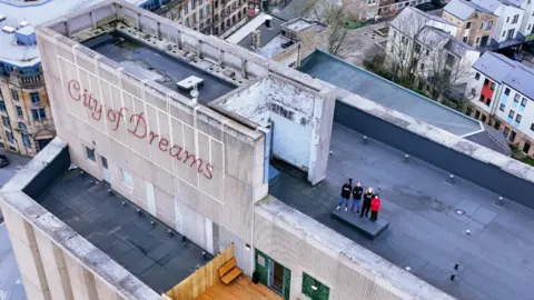 Other An aerial view of a large concrete building with red writing on the side which says 'City of Dreams'. There are four people stood on the large flat roof of the building. 