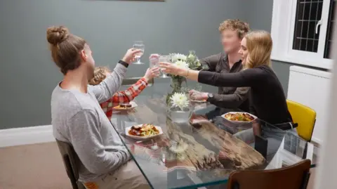 S4C The image shows three adults and one child seated around a rectangular glass dining table inside a room with grey walls. Plates of food are on the table, along with drinking glasses and a vase containing white flowers. All visible people have their arms raised, holding glasses toward the centre of the table. Chairs of different styles surround the table, and a radiator and window are visible on the wall behind them.