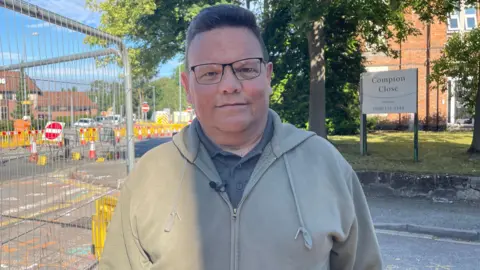 BBC A man with short black hair is wearing glasses, a light green hooded jumper and a grey shirt. He is standing next to a metal barrier besides roadworks. 