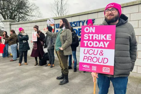 A picket line at University of Aberdeen with a striker holding up a cardboard sign that reads: "On strike today / UCU & proud".