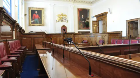 Inside the Guernsey States Chamber. Paintings in frames and a crest are on the wall while microphones sit in front of seats lining the sides and throughout the room.