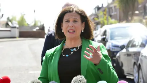 PAcemaker Sinn Féin leader Mary Lou McDonald, pictured outside on a sunny day, standing in front of a microphone and wearing a green jacket, while gesturing with her hand