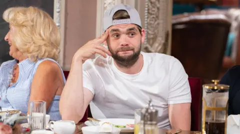 BBC/Studio Lambert/Euan Cherry Adam, sitting at a table, with food in front of him, wearing a white T-shirt, and cap on his head, with it turned round the other way. He has a dark beard and dark hair, peaking through the front of the cap. Fiona is sat to his right wearing a blue and white stripped short-sleeved top with long fair hair. Her face is to the left. 