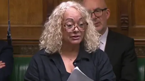 Parliament TV Kim Johnson, who has curled blonde hair and is wearing glasses, is standing up in the House of Commons, holding a speech in front of her, in a scene from Parliament TV. 