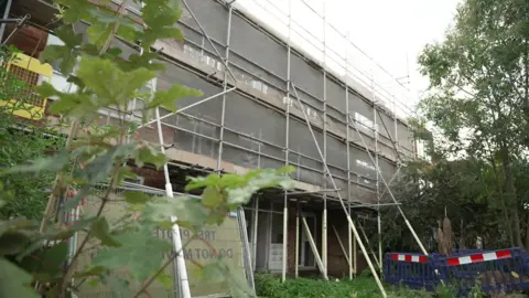 Scaffolding surrounds the condemed block of flats, with barriers to prevent access in the overgrown garden. 