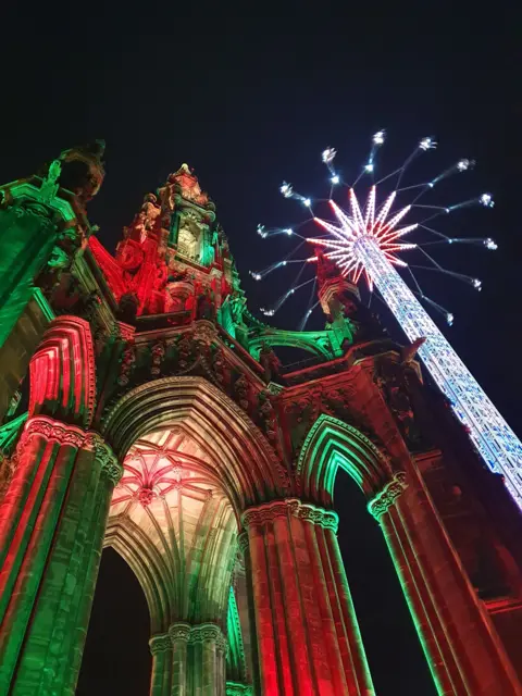 Carolyn Richardson Portrait image of the Scott monument in Princes Street Gardens, Edinburgh from below, illuminated in red and green lights and a fair ride next to it with swings at the very top, illuminated in bright lights against a dark sky