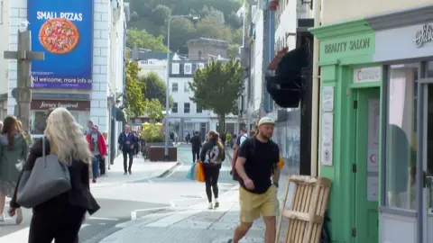 A woman wearing a grey handbag on the right walking down Broad Street in St Helier. There is a man wearing yellow shorts, a black t-shirt and a cap walking past a shop which is green. There are other people in the distance walking up and down Broad Street. The pavement is light grey and there are buildings in the background