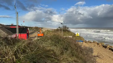 Hemsby beach and lifeboat station, with a digger on the beach repositioning concrete sea defence blocks. The lifeboat station is set back from the beach. It is a red building. The sea is at high tide, with white surf on the water.
