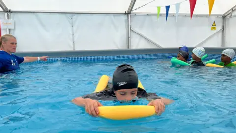 A swimming instructor in a blue shirt guides children using colourful foam noodles in an indoor pool, beneath a white canopy with multicoloured bunting overhead.