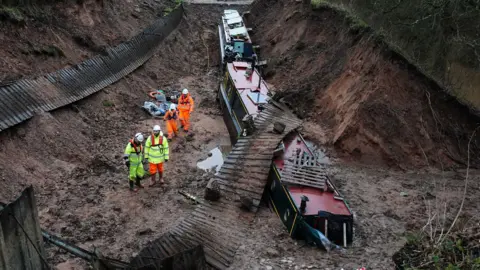 Two boats at the bottom of a hole in a canal with no water in. one is sunk partially into the earth and has a wooden fence lying over the top of it. there are four people standing in the hole next to them in fluorescent yellow and orange clothing with white hard hats