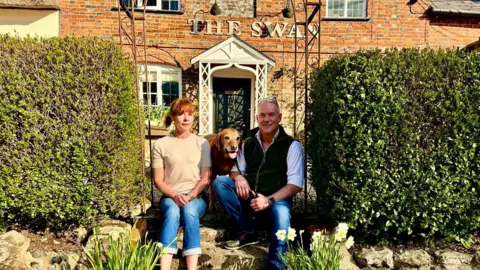 Geraldine Symonds A husband and wife - Sophie Cooper and Andrew Wiggins - sitting with a large golden-coloured dog outside the front of the Swan pub with green bushes and yellow daffodils either side of them and the 16th century thatched pub in the background. 
