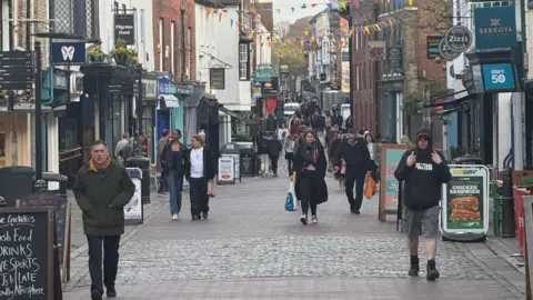 Keir Mackenzie/BBC Canterbury High Street, with multiple old buildings housing shops on either side. It is a bright evening and there are a modest amount of pedestrians walking down the road.