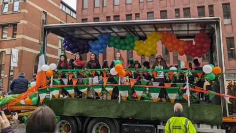 A truck float with a group of female Irish dancers surrounded by green, white and orange flags and multi-coloured balloons. 