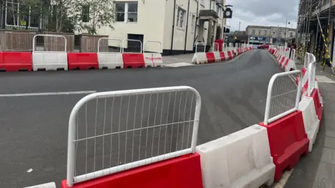 The high street road running through Cane with red and white barriers on either side and buildings on each side of the road.