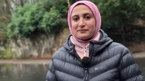 Sondos is standing in a park, there is a lake behind her. She is wearing a pink headscarf, pink jumper and a black jacket. 