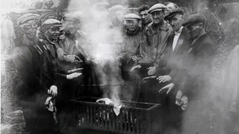 Hulton Archive/Getty Images Workers in flat caps are warming their hands around a brazier during the General Strike.