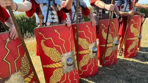 A line of bright red Roman recreation shields, with gold centres and yellow markings on them. They are held by people dressed as Roman soldiers. 