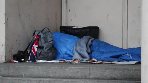 A person is sleeping in a blue sleeping bag in a door way. Next to them is a backpack.