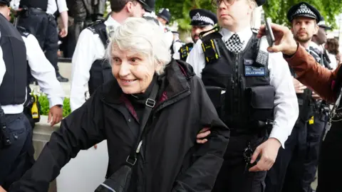 Jeff Moore Reverend Sue Parfitt with two police officers either side of her. She is wearing a vicar's collar and a back coat while sat near white stones.