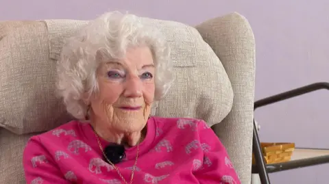 Nora Hoskin, sat on a cream armchair, she is weaing a pink jumper with a gold necklace, she is facing the camera directly. The wall behind her is purple. 