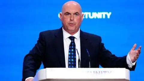Paul Davies standing behind a white lectern giving a speech at a Conservative conference. The word opportunity is partly visible, the background behind him is bright blue.