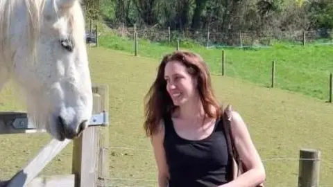 Belinda Taylor is stood wearing a black sleeveless vest with long brown hair. She is stood by a wooden gate with a white horse on the other side.