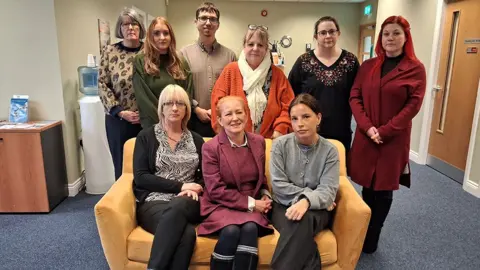 LDRS The staff of Lincolnshire Centre for Grief and Loss pose for the camera in their office. Three women sit on a yellow sofa with six people standing behind them. 
