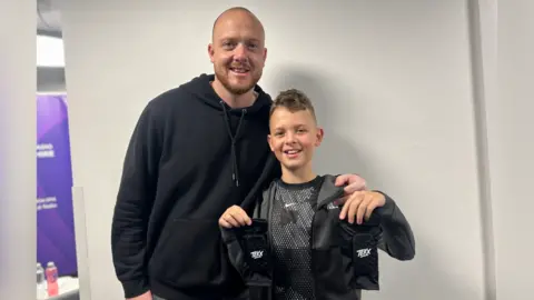 Ashton has short hair and is wearing a jumper and black top, while Sam, who has a beard and a shaved head, is wearing a black hooded top.