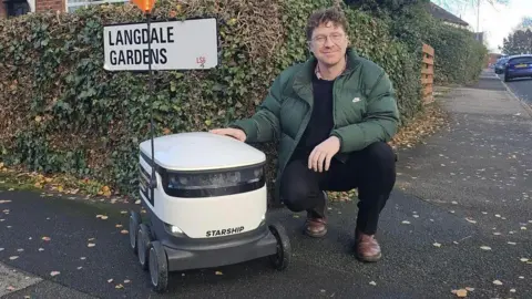 Councillor Jonathan Pryor poses for a photo with a delivery robot. The machine has six wheels and is around 60cm in height. Councillor Pryor has curly strawberry blonde hair and glasses.