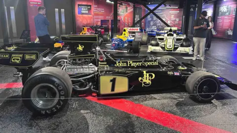 Kate Bradbrook/BBC A black racing car with gold writing that says "John Player Special", with a intertwined letter JPS logo and a number one in black with a gold background.
It has large Goodyear tyres. It has Ronnie Peterson's name on the body about where the driver's seat begins. It is parked on a black stage at an exhibition.