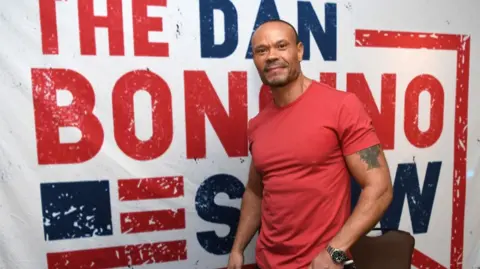 Dan Bongino, wearing a red T-shirt, stands in front of a big red white and blue sign saying The Dan Bongino Show