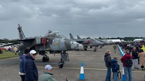 BBC A number of fighter jets lined up in a taped off area on an airfield. People have gathered around to look at the aircraft.