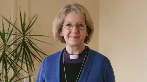 Right Reverend Jackie Searle wearing her vicar's collar, purple tunic, black top and blue cardigan stood in front of a leafy plant.