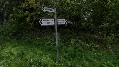 Google The picture shows a rural road junction marked by three directional road signs standing on a grassy roadside. The arrows point toward Lealholm, Glaisdale and Rosedale, and Danby and Fryup. Behind the signs is dense greenery with trees and bushes.