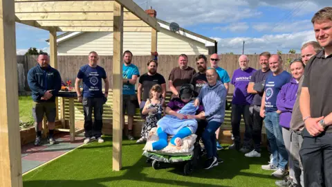 A group of people standing in an astroturf area smiling and looking at the camera. They are surrounding a family of 4, 2 adults and 2 children, one of which is Lucas who is in his wheelchair in the garden.