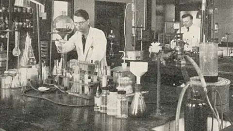 Yorkshire Cancer Research A black and white photo of two men in white lab coats working in a research lab in Leeds. There are lots of flasks and experiments going on on the desks in front of them.