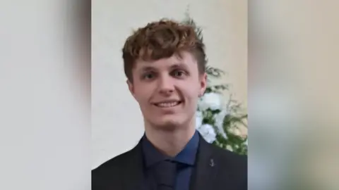 Family handout A still image of Kacper Dudek. He is a young man with short, light brown hair and is wearing a black suit jacket, a black tie and a navy shirt. He is standing in front of a bouquet of white flowers.