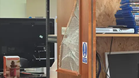 Age UK Milton Keynes A broken window, in a charity show, showing DVDs on shelves to the right, a computer monitor to the left and right, and a hot chocolate machine in the desk.