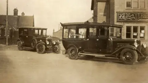 Rebecca Hillier A black and white photo of the funeral service building on the right with H.Hillier written on the exterior. There are two very old cars from the 1920s parked outside on the empty road.