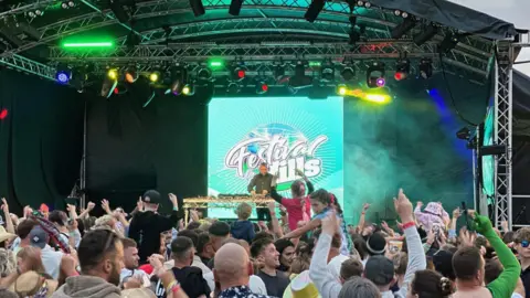 Image of a festival stage with a DJ standing in front of a Festival on the Hills banner, with a crowd of festival goers watching him and dancing