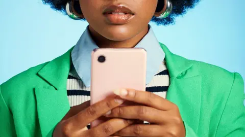 A woman looking at her smartphone. she is wearing a green jacket, blue shirt and blue and white stripe top and is in front of a plain blue background
