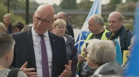 Bald man in a suit and glasses speaking to a small group of people outdoors, with others standing behind holding flags and signs near a riverside setting
