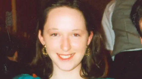 Josephine Dullard smiles at the camera, she has shoulder length brown hair and gold hoop earrings. She is wearing a blue dress.